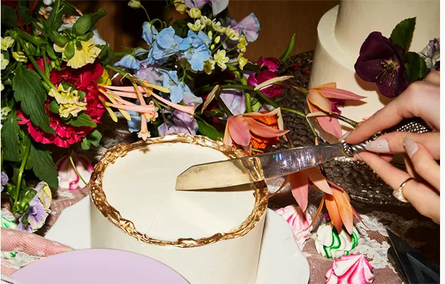 A hand cutting a celebration cake set among vivid seasonal flowers