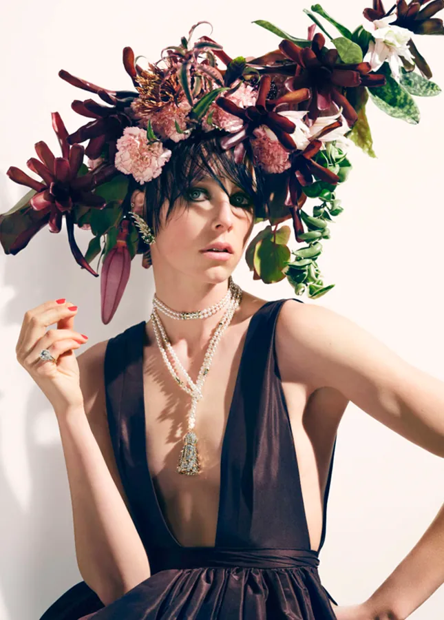 A model in a dark gown beneath a jewel-toned floral headdress and pearl necklace