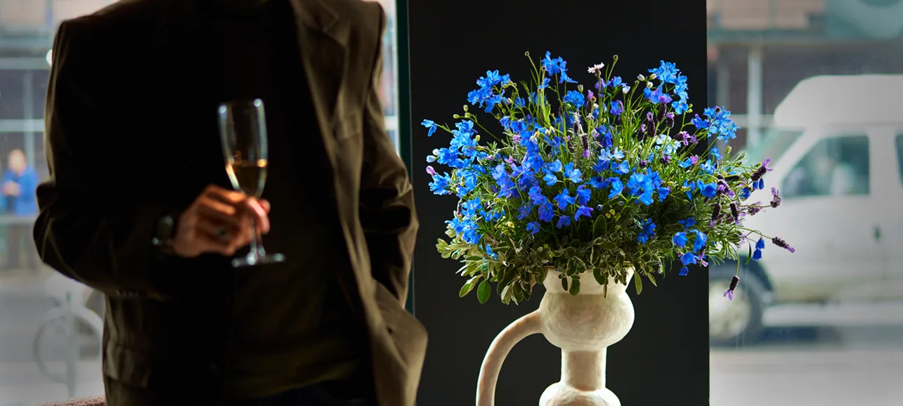 A guest in a dark blazer holding a champagne flute beside a sculptural white vase of blue delphinium, styled by Buunch for an event