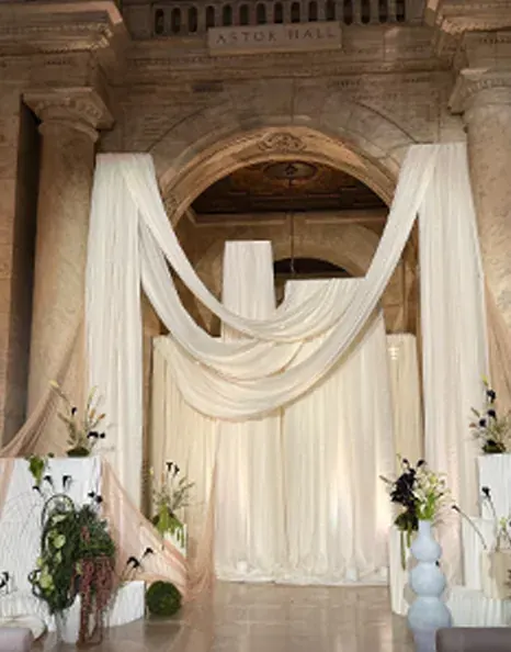 White fabric draped beneath a stone archway with sculptural floral arrangements on plinths.