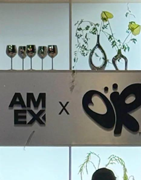 A branded wall reading “AM EX × OR” with wine glasses and two bud vases