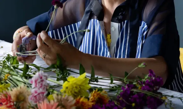 A floral designer in a striped apron arranging yellow, pink and orange blooms on a white surface.