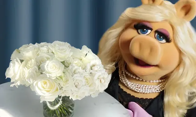 Miss Piggy in a pearl necklace beside a glass vase of white roses in front of a blue curtain.