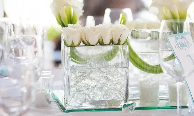 A glass vase of white roses filled with clear beads on a mirrored tray among wine glasses.