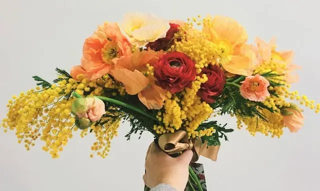 A hand holding a bridal bouquet of orange poppies, red ranunculus and yellow mimosa.