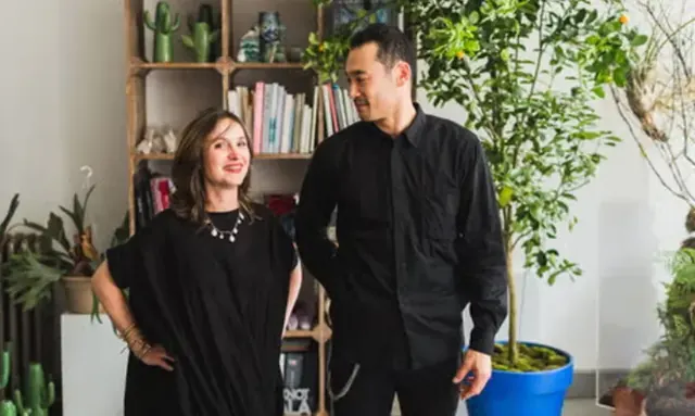 A woman in a black dress and a man in a black shirt smiling in a bright studio with plants.