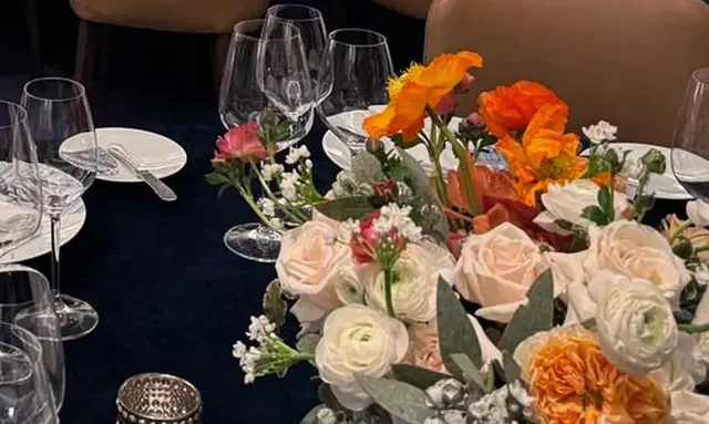 A low centerpiece of orange and blush roses with greenery on a dark set table.