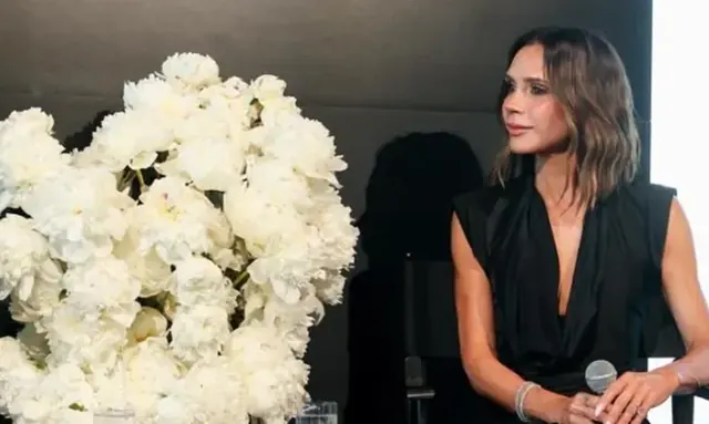 Victoria Beckham holding a microphone beside a large arrangement of white flowers.