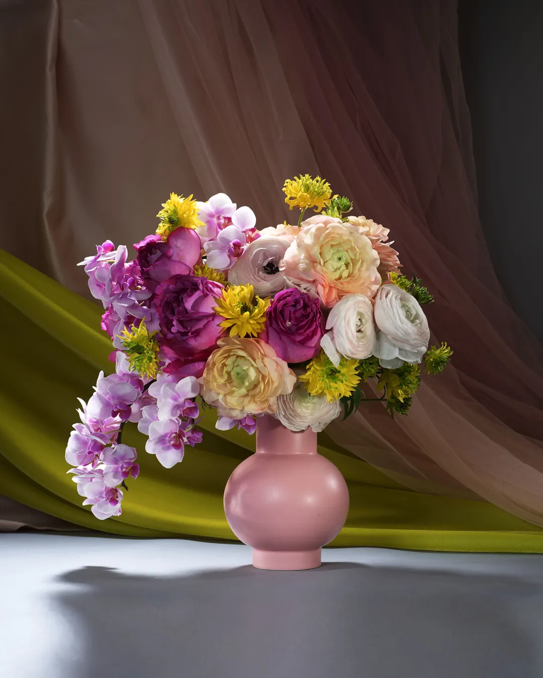 Pastel ranunculus, fuchsia roses and cascading orchids arranged in a blush-toned vase