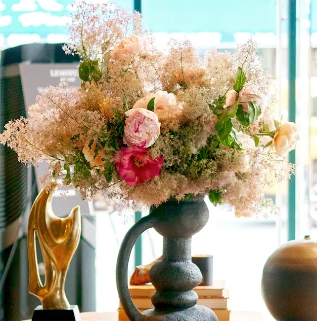 Blush peonies and airy pale pink flowers arranged in a dark sculptural vase in a bright interior.